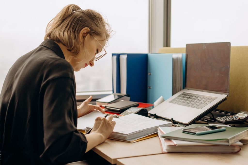 Frau mit Brille arbeitet am Schreibtisch Unterlagen durch, daneben Ordner, ein Laptop und ein Taschenrechner